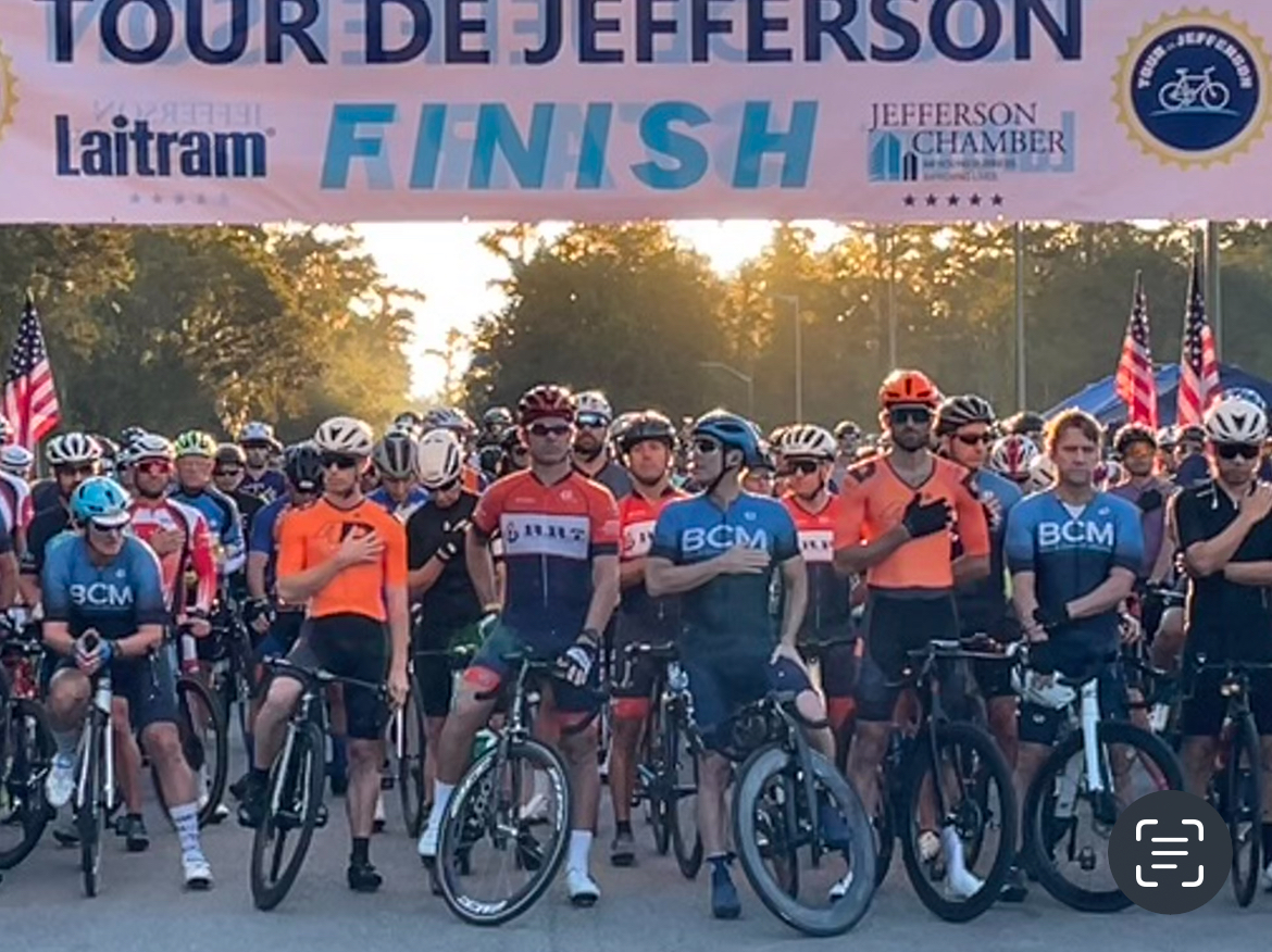 Cyclists gather at the starting line under a banner for the Tour de Jefferson event. Some wear matching jerseys. Bicycles and helmets are visible.